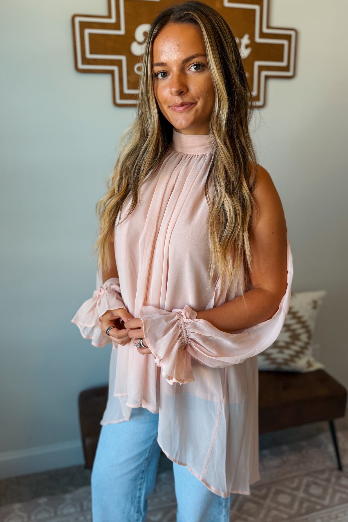A woman wearing a pale blush pink high-neck blouse with dramatic gathered sleeves and light blue jeans, standing in a home interior setting.