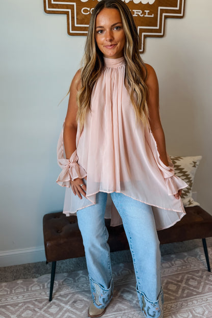 A woman wearing a pale pink sleeveless high-neck blouse with ruffled sleeves and a draped silhouette, paired with light blue jeans and western boots, standing in a home setting.