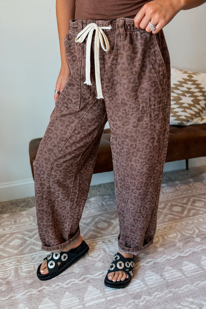 A person wearing loose brown leopard-print pants with a cream drawstring, standing indoors on a patterned rug near a bench.