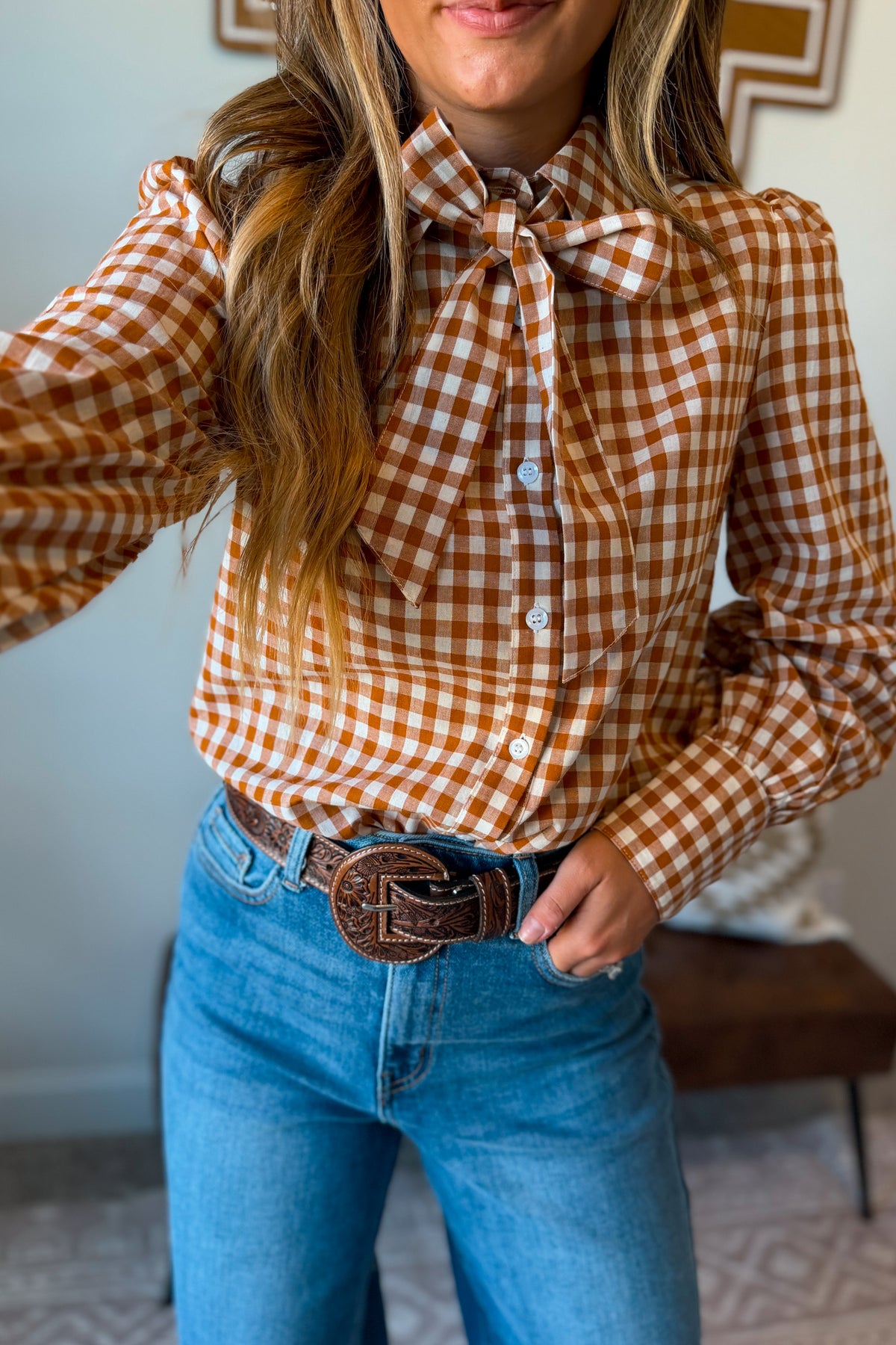 A woman wearing a long-sleeve brown and white gingham bow tie blouse tucked into blue jeans with a brown leather belt.
