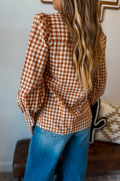 A woman wearing a rust-orange and white gingham long-sleeve blouse with blue jeans, shown from a back-three-quarter angle in an indoor setting.