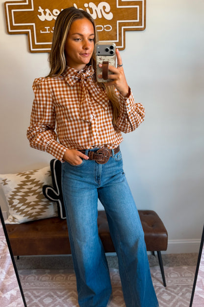 A woman takes a mirror selfie wearing a rust-orange and white gingham long-sleeve blouse with a bow tie collar, paired with high-waisted blue flare jeans and a brown leather belt with a large buckle.