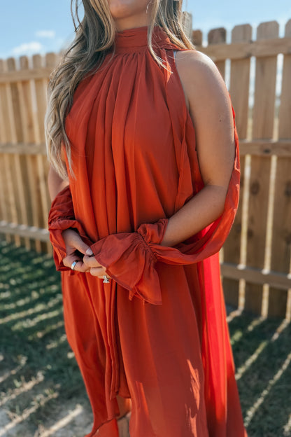 A woman wearing a sleeveless rust-orange dress with a high gathered neckline and long sheer sleeves tied at the wrists, standing outdoors in front of a wooden fence.