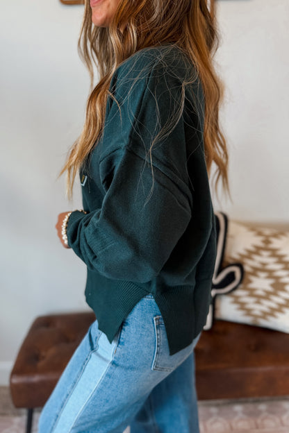 A woman wearing a dark forest green knit sweater and light blue jeans, shown from a side-back angle in an indoor setting.