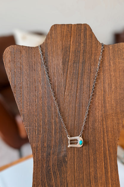 A silver-toned necklace with a buckle shape pendant and a small turquoise accent, displayed on a wooden mannequin.