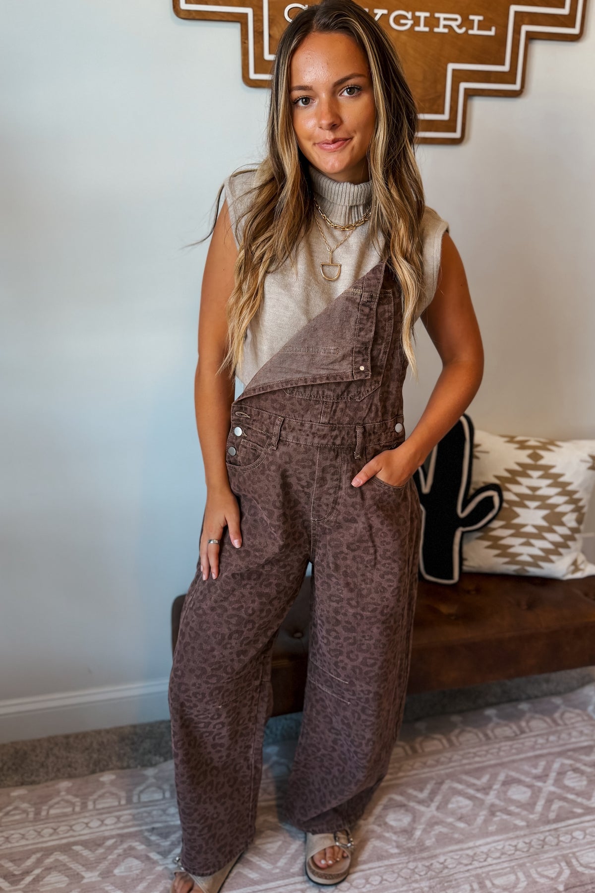 A woman modeling brown leopard-print wide-leg overalls over a cream sleeveless top.