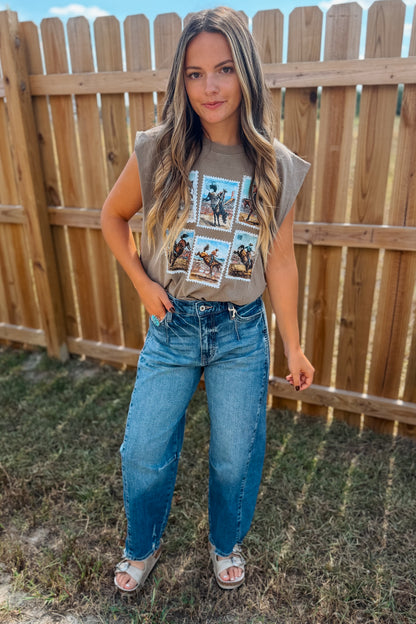 A woman stands outdoors in front of a wooden fence wearing a sleeveless taupe graphic t-shirt and blue jeans, and white slide sandals.