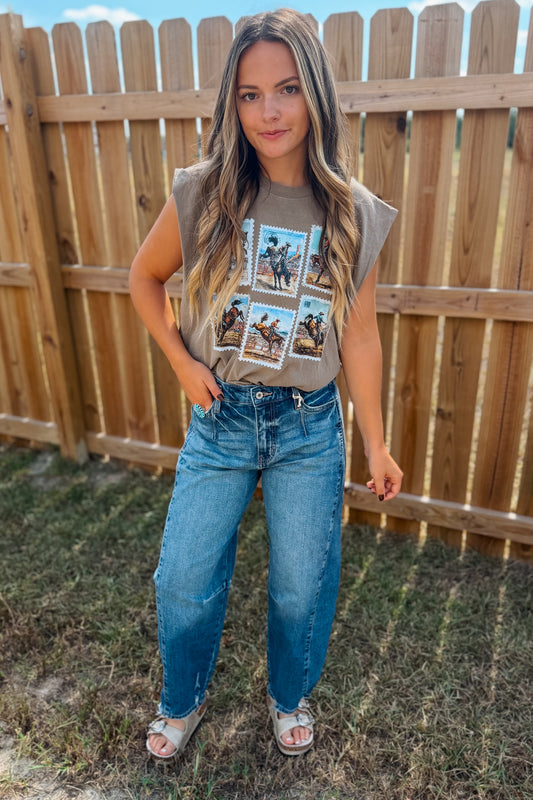 A woman stands outdoors in front of a wooden fence wearing a sleeveless taupe graphic t-shirt and blue jeans, and white slide sandals.