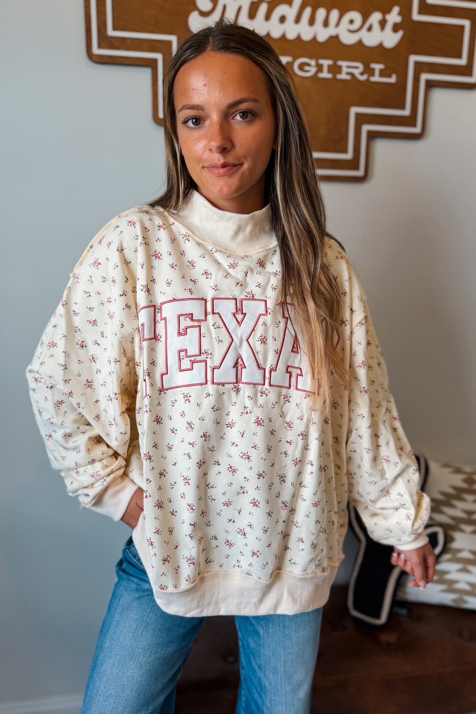 A woman wearing a cream-colored crewneck sweatshirt with a small floral print and 'TEXAS' outlined in red across the chest, paired with blue jeans.