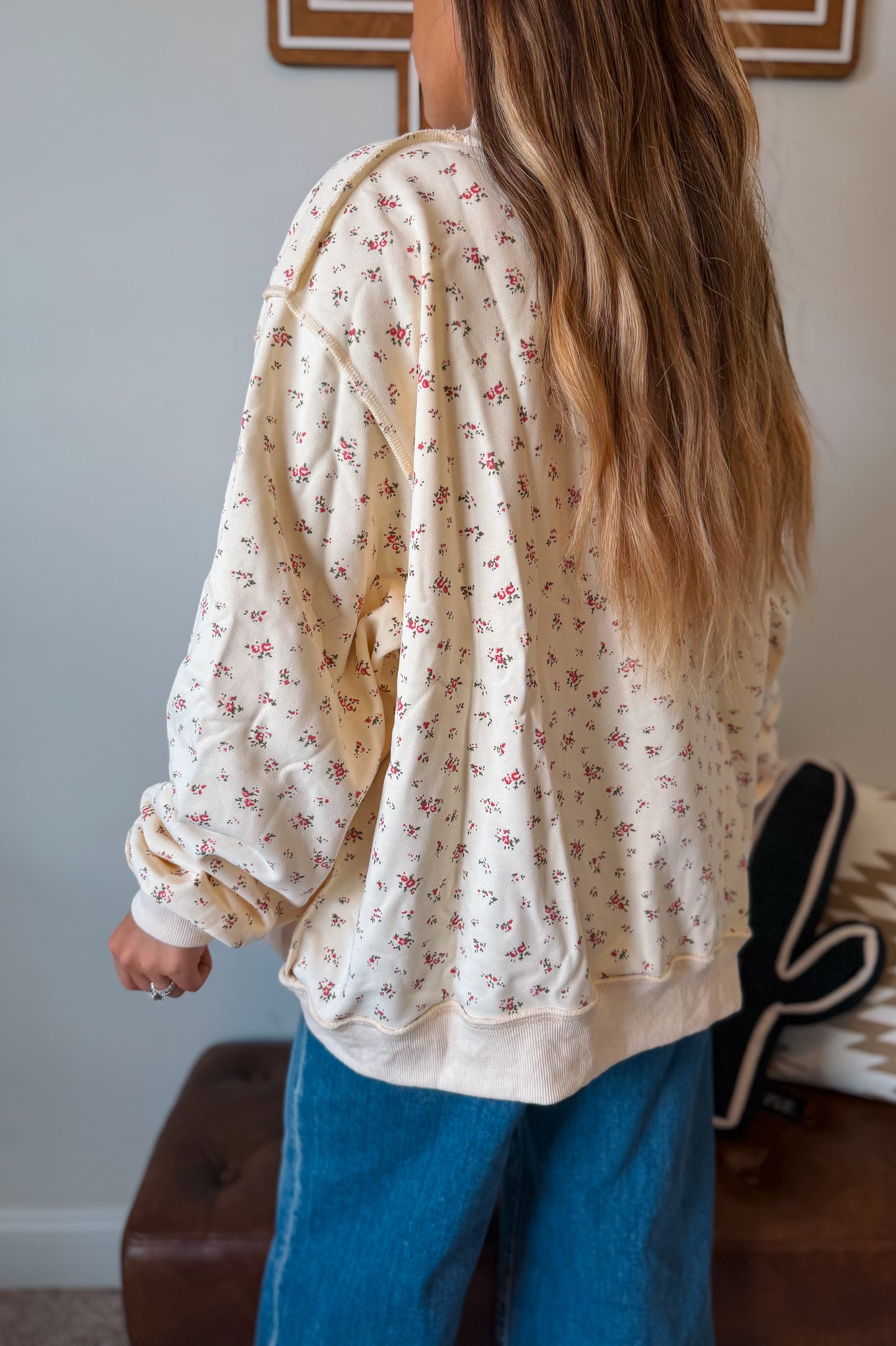 A woman shown from behind wearing a cream-colored, oversized long-sleeve sweatshirt with a small pink floral print, paired with blue jeans in an indoor setting.