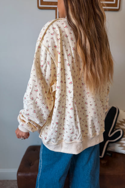 A woman shown from behind wearing a cream-colored, oversized long-sleeve sweatshirt with a small pink floral print, paired with blue jeans in an indoor setting.