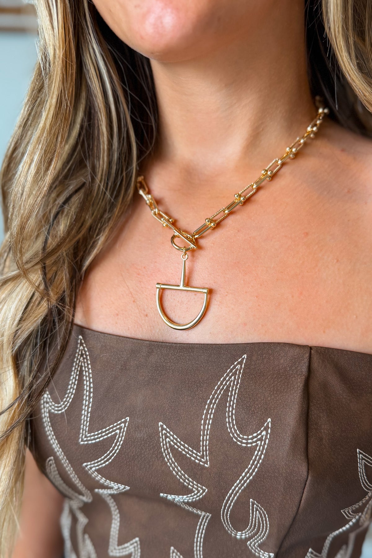 A close-up of a model wearing a gold chain necklace with a unique open D-shaped pendant, paired with a brown off-the-shoulder embellished top.