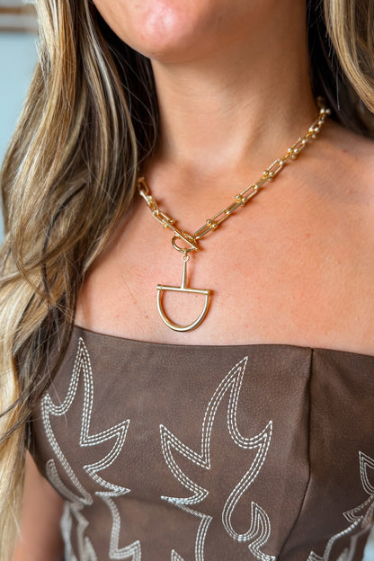 A close-up of a model wearing a gold chain necklace with a unique open D-shaped pendant, paired with a brown off-the-shoulder embellished top.
