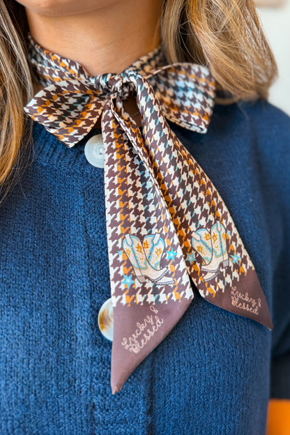 A close-up of a person wearing a patterned silk necktie styled as a bow at the collar of a blue knit sweater. The tie features a brown, beige, and orange houndstooth pattern with embroidered blue and orange hearts and the words 'Love & Bliss' on the pointed ends.
