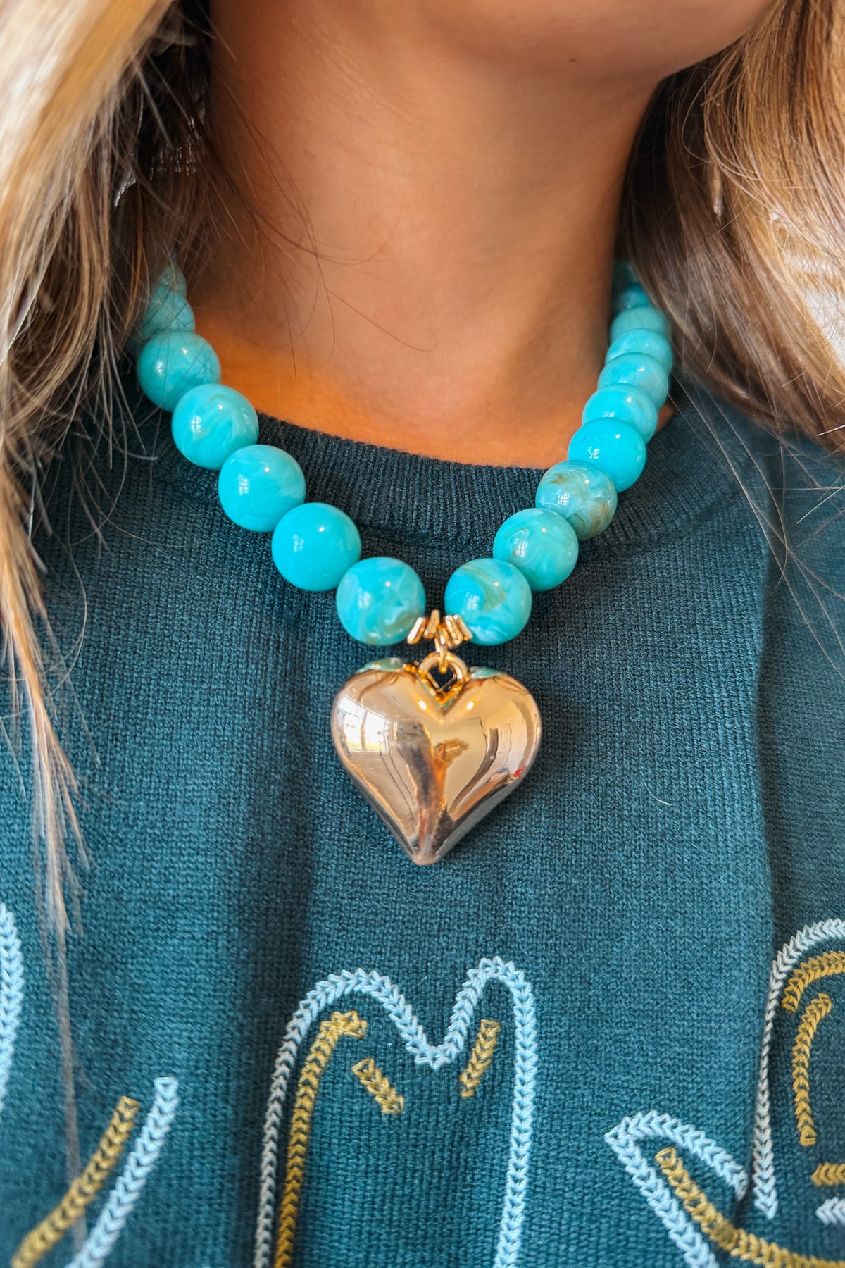 A close-up of a person wearing a turquoise beaded necklace with a polished gold heart pendant, paired with a dark teal sweater.