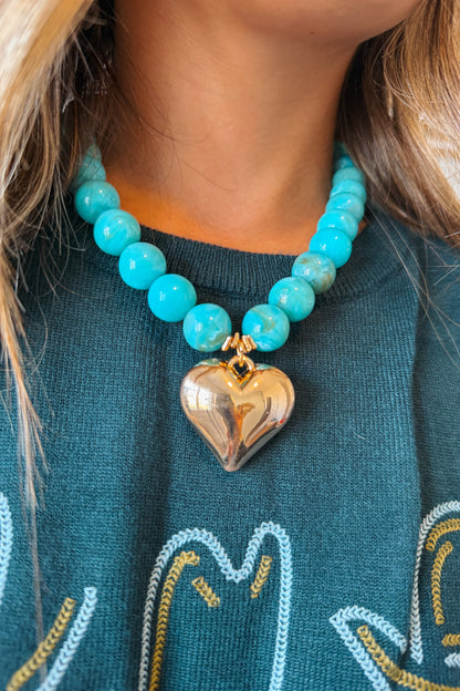 A close-up of a person wearing a turquoise beaded necklace with a polished gold heart pendant, paired with a dark teal sweater.