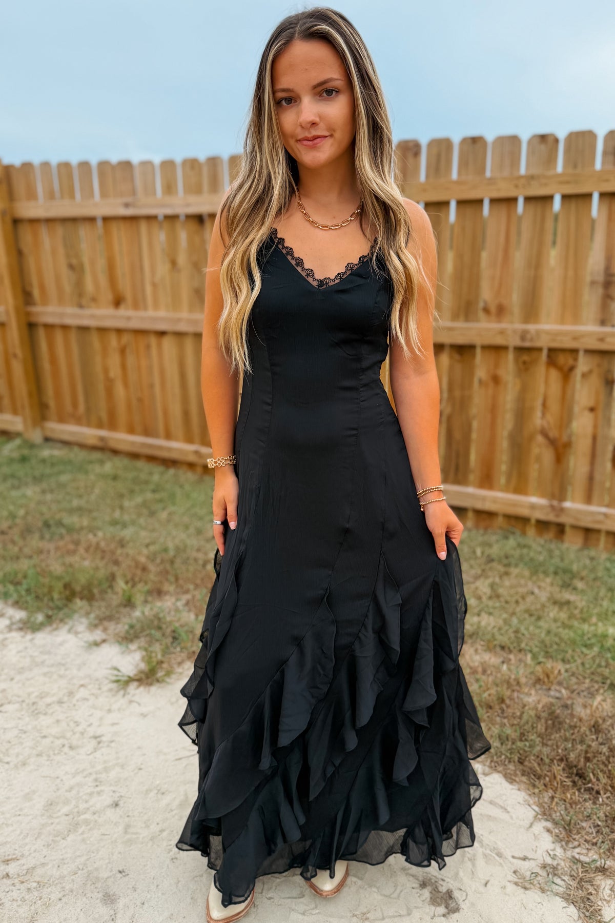 A woman wearing a black sleeveless maxi dress with a ruffled tiered skirt, standing outdoors in front of a wooden fence.