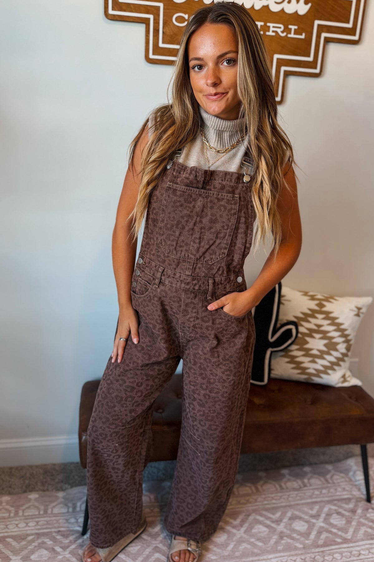 A woman modeling brown leopard-print wide-leg overalls over a white turtleneck, standing in a room with a patterned rug and a bench.