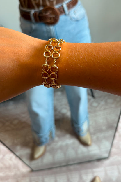 A gold-toned multi-strand link bracelet worn on a person's wrist, with light blue jeans, a brown belt, and beige boots visible in a mirror reflection.
