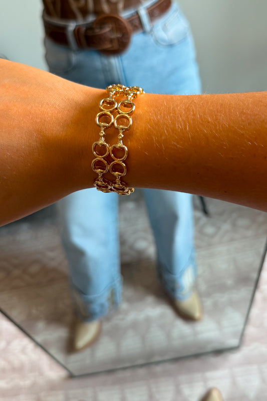 A gold-toned multi-strand link bracelet worn on a person's wrist, with light blue jeans, a brown belt, and beige boots visible in a mirror reflection.