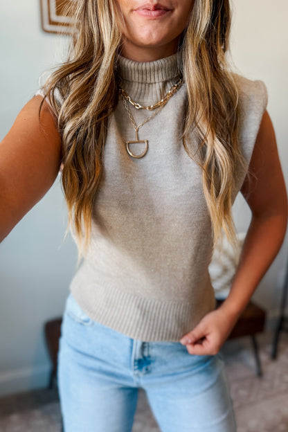 A woman wearing a sleeveless ribbed knit turtleneck sweater in a light oatmeal beige color, paired with light blue jeans and layered gold necklaces.