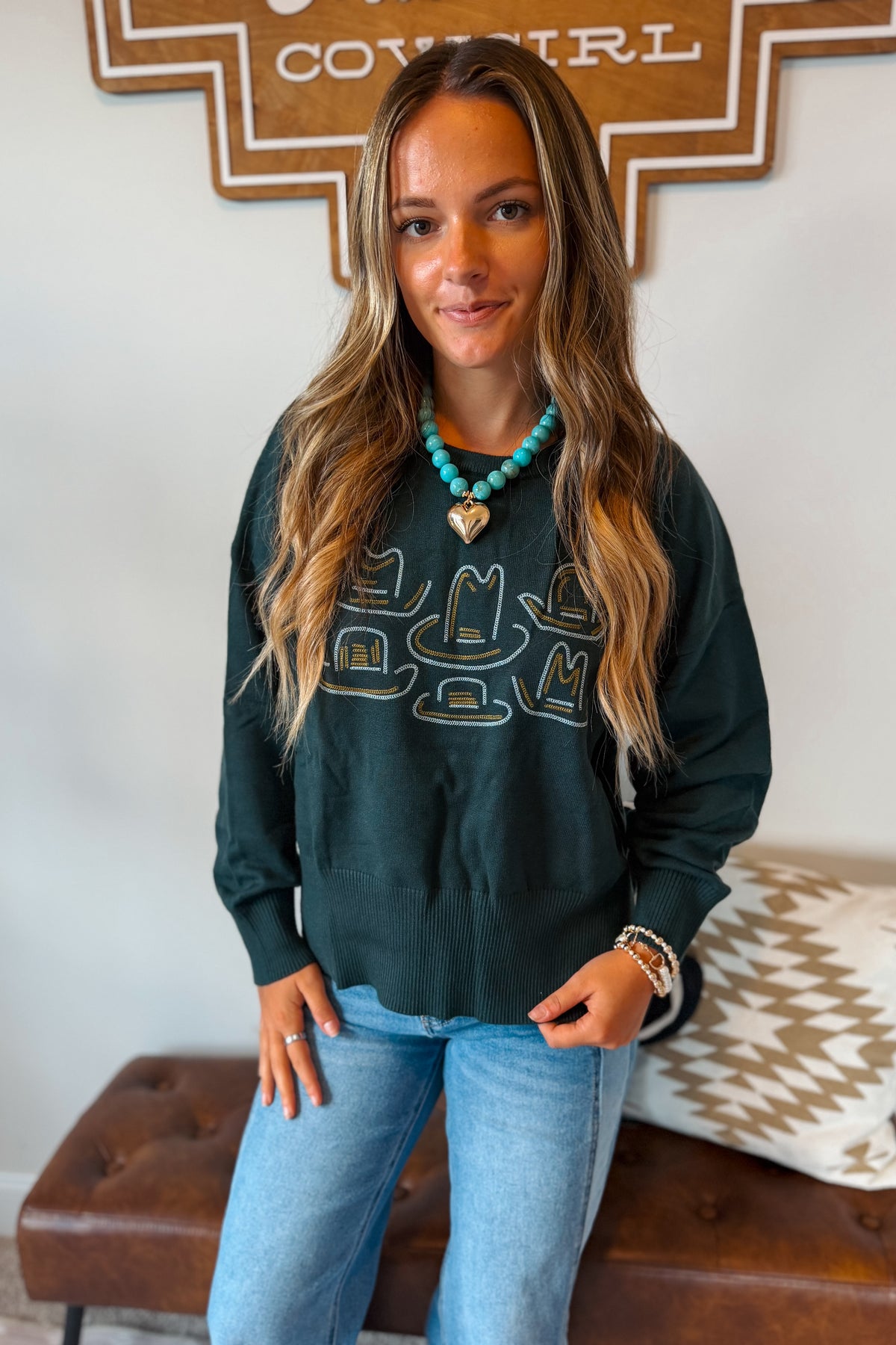A woman wearing a dark green long-sleeve sweatshirt with a graphic of cowboy hats in gold and turquoise, paired with light blue jeans and a turquoise necklace.