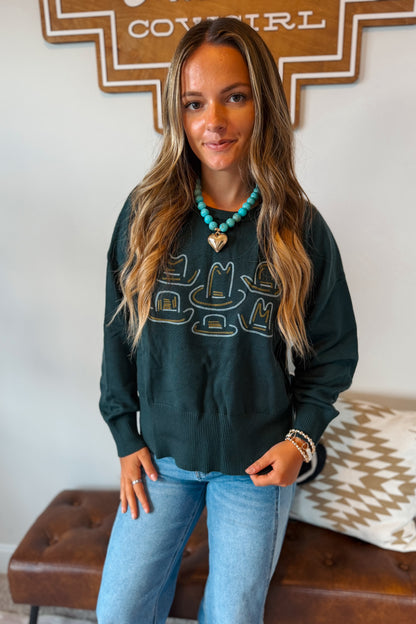 A woman wearing a dark green long-sleeve sweatshirt with a graphic of cowboy hats in gold and turquoise, paired with light blue jeans and a turquoise necklace.