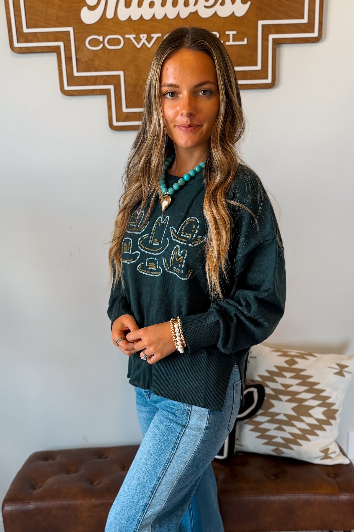 A woman modeling a dark green long-sleeve graphic sweatshirt with turquoise and white lettering, paired with light blue jeans, standing in front of a white wall with a brown sign and a patterned pillow.