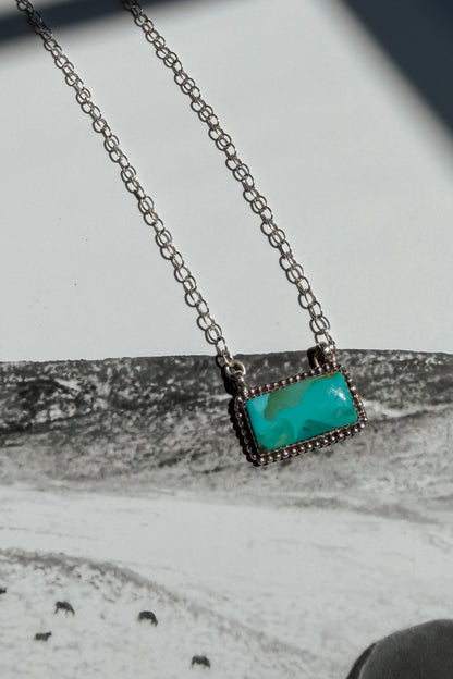 A silver-toned chain necklace with a rectangular turquoise pendant, displayed on a textured gray surface.