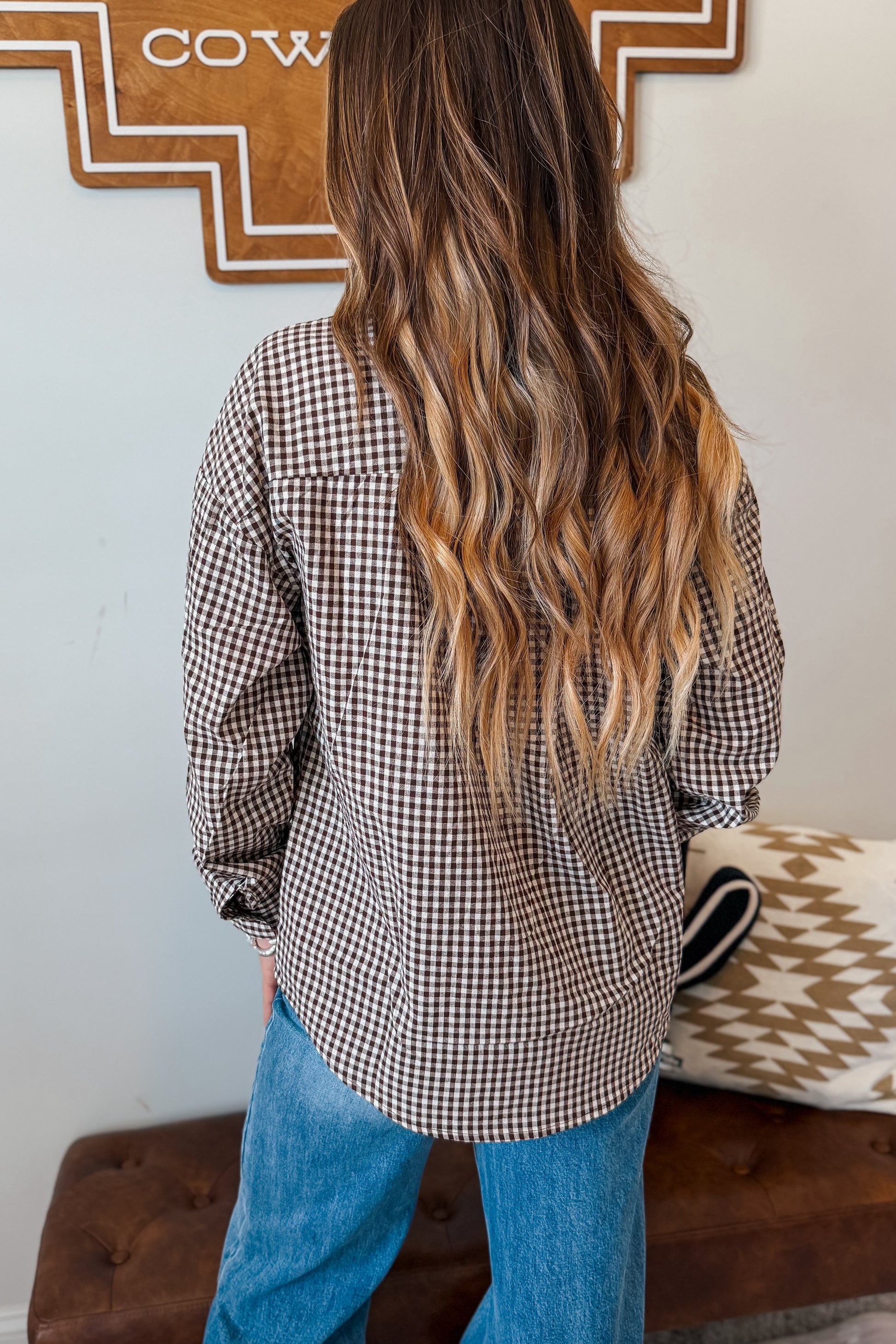 A woman viewed from behind wearing a brown and white and brown gingham long-sleeve button-up shirt and blue jeans