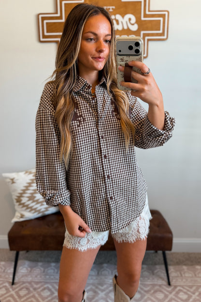A woman wearing a brown and white gingham button-up shirt with lace shorts and cowboy boots