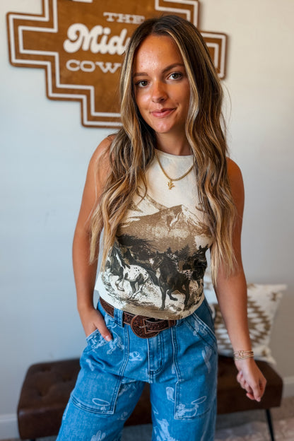 A woman modeling a sleeveless graphic tank top with a western mountain and horse print, paired with blue jeans and a brown leather belt, standing in front of a wall with a wooden sign.