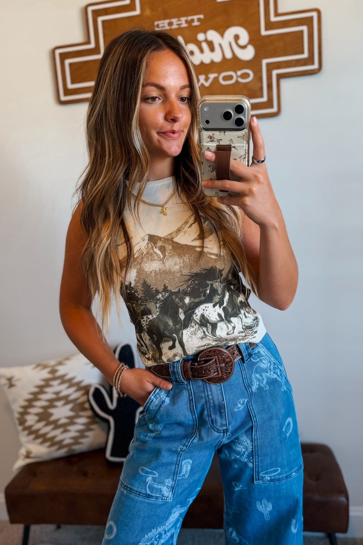 A woman taking a mirror selfie wearing a beige and black graphic print tank top and high-waisted blue denim jeans with a brown leather belt.