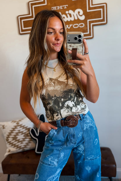 A woman taking a mirror selfie wearing a beige and black graphic print tank top and high-waisted blue denim jeans with a brown leather belt.