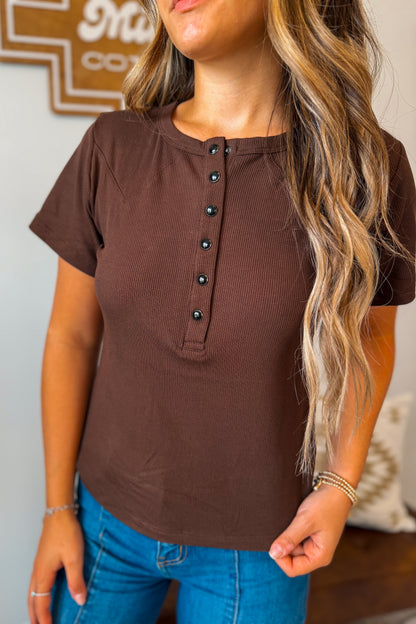 Brown ribbed knit top featuring a quarter-button neckline, short sleeves, and a fitted silhouette. Designed with a soft textured fabric for comfortable everyday wear.