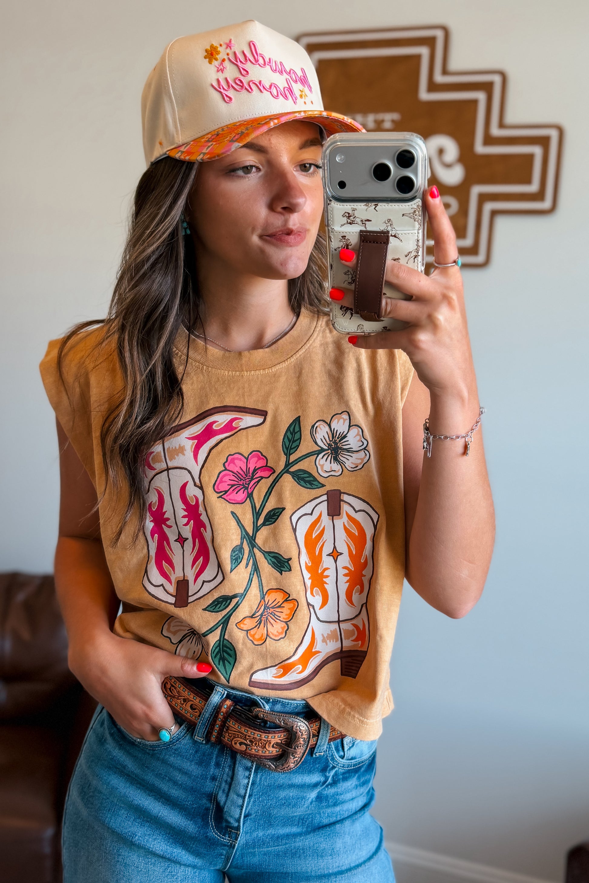 Person wearing a tan cap and graphic t-shirt, taking a mirror selfie.
