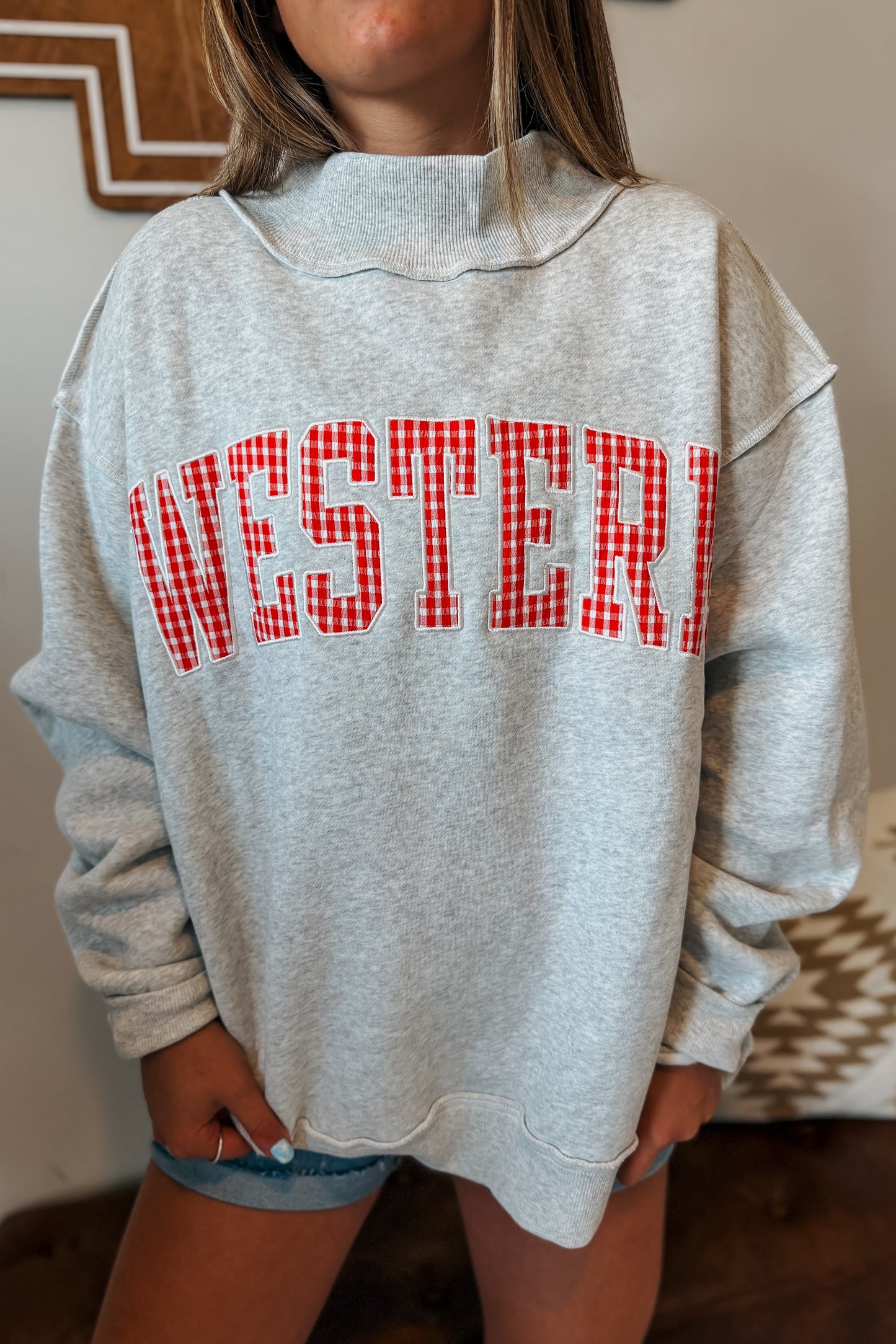 Close-up of “WESTERN” embroidery with red gingham plaid fabric inlay on a gray mock neck sweatshirt.