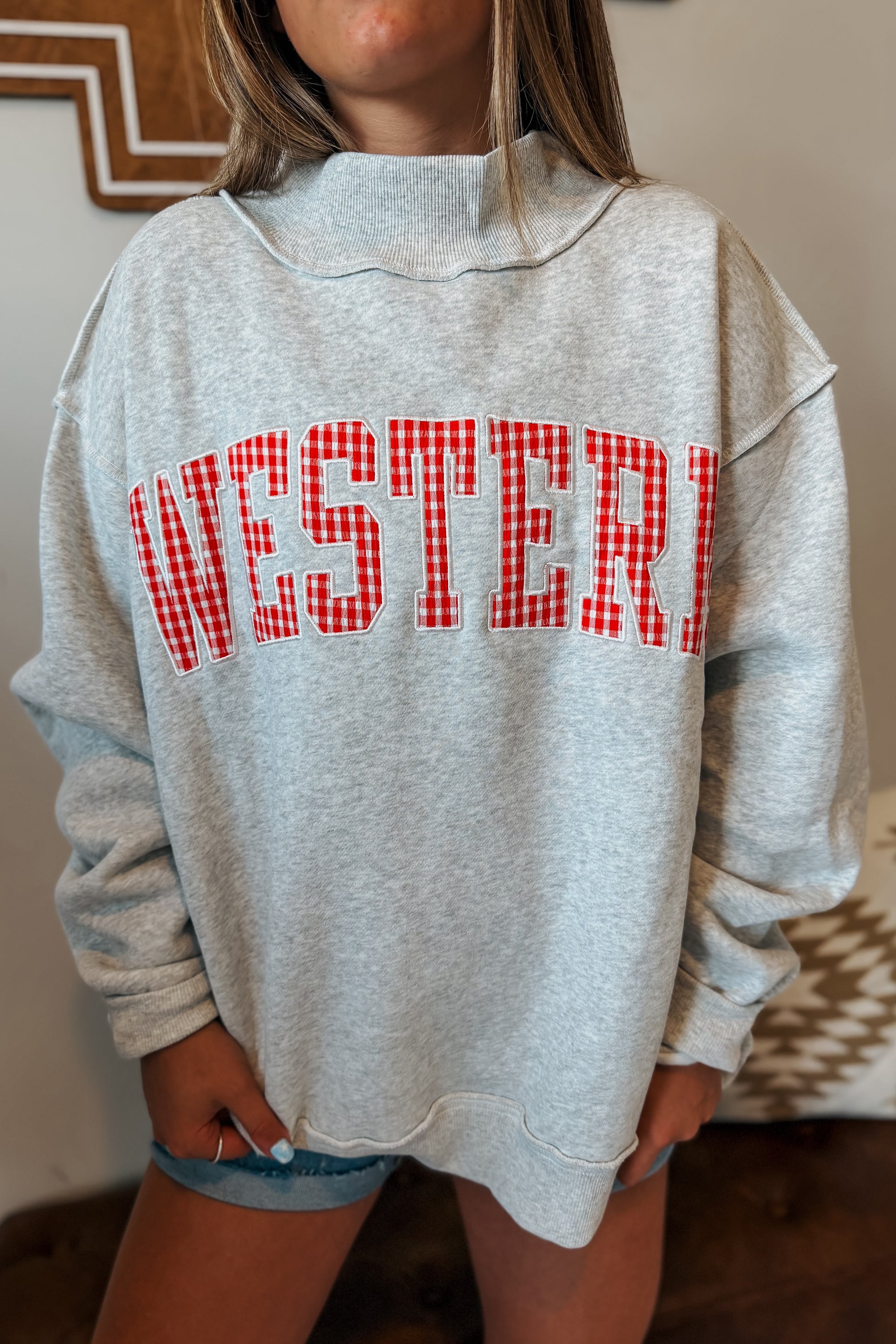 Close-up of “WESTERN” embroidery with red gingham plaid fabric inlay on a gray mock neck sweatshirt.