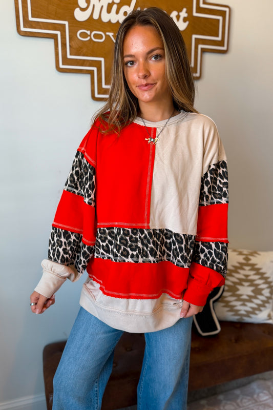 A woman wearing a color-blocked long-sleeve sweatshirt with red, cream, and leopard print panels, paired with blue jeans, standing in an indoor setting.