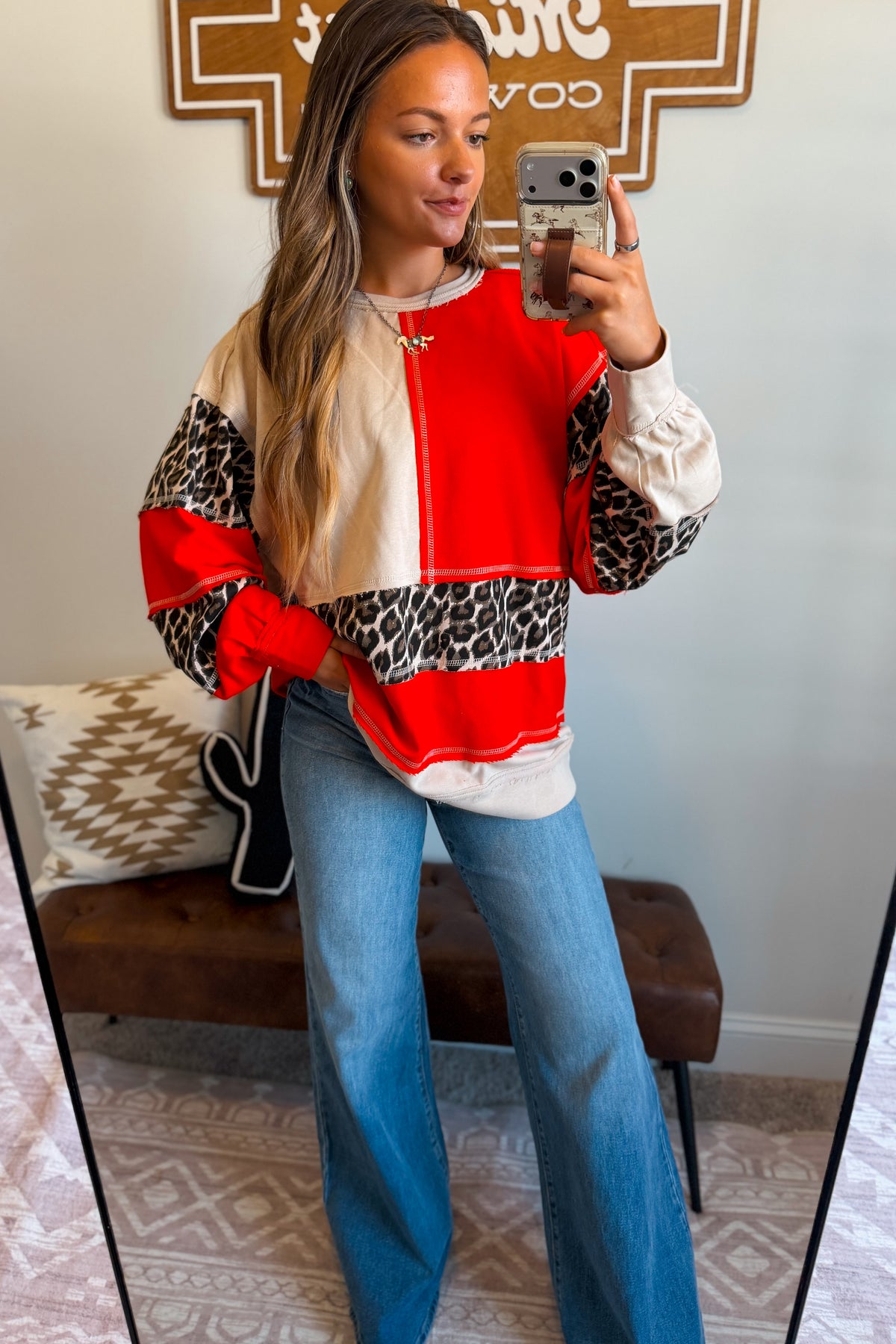 A woman takes a mirror selfie wearing a color-blocked sweatshirt with red, cream, and leopard print panels, paired with blue jeans in a home setting.