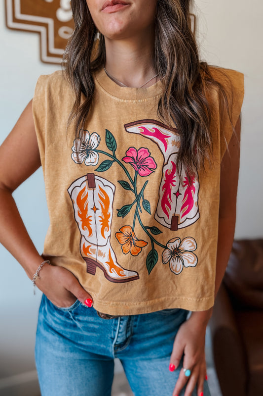 Person wearing a tan sleeveless shirt with cowboy boot and floral design, paired with blue jeans.