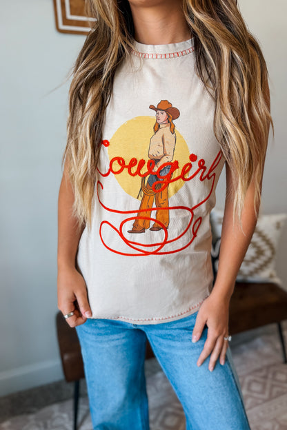 A woman wearing a tan-colored graphic tank top with a western 'cowgirl' design featuring a cowboy illustration and red script, 