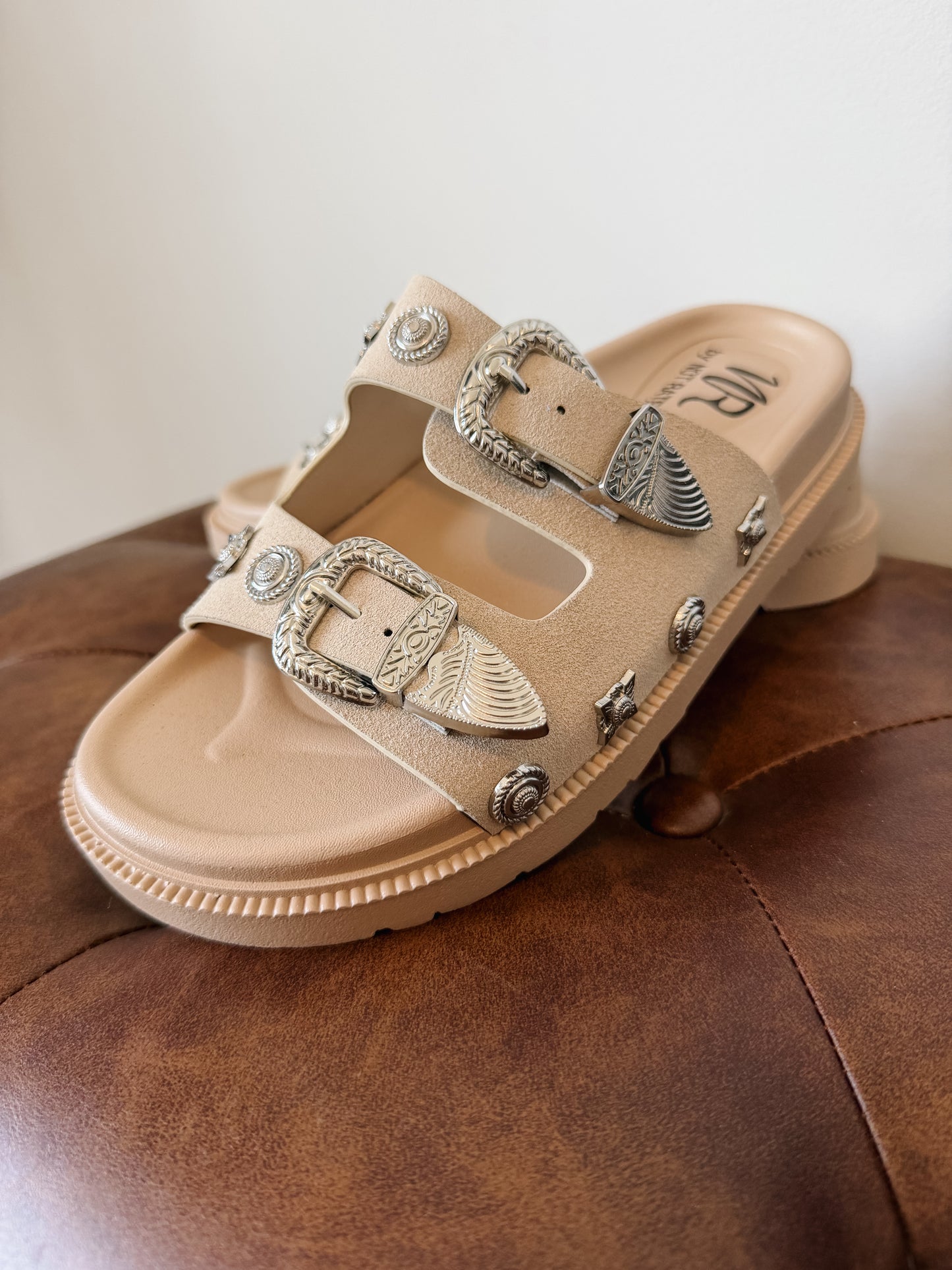 Cream western slide sandals featuring engraved buckle accents, metal stud detailing, and a cushioned footbed.