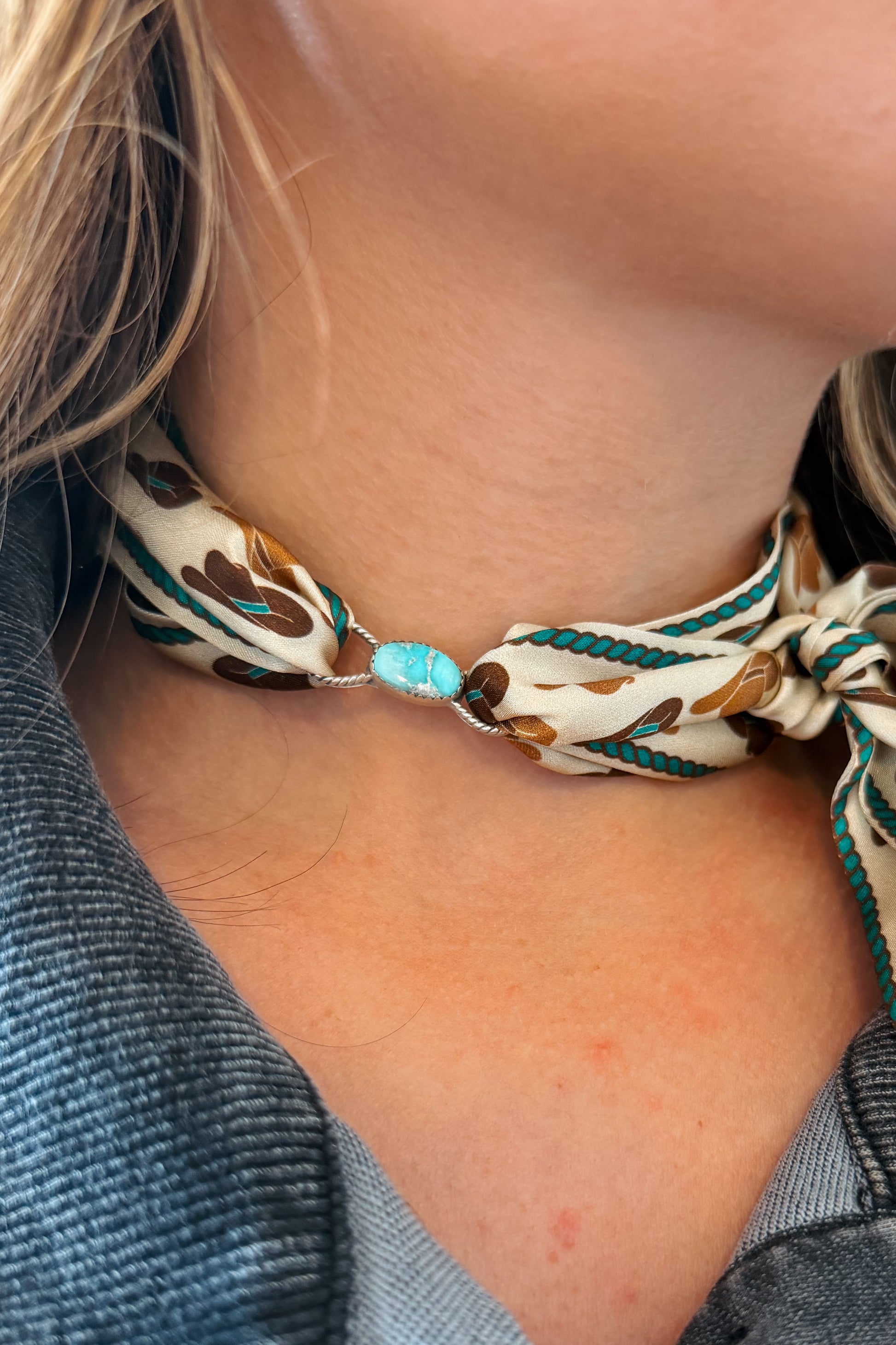 A close-up of a person's neck wearing a patterned silk scarf tied around the collarbone, featuring a turquoise stone centerpiece.