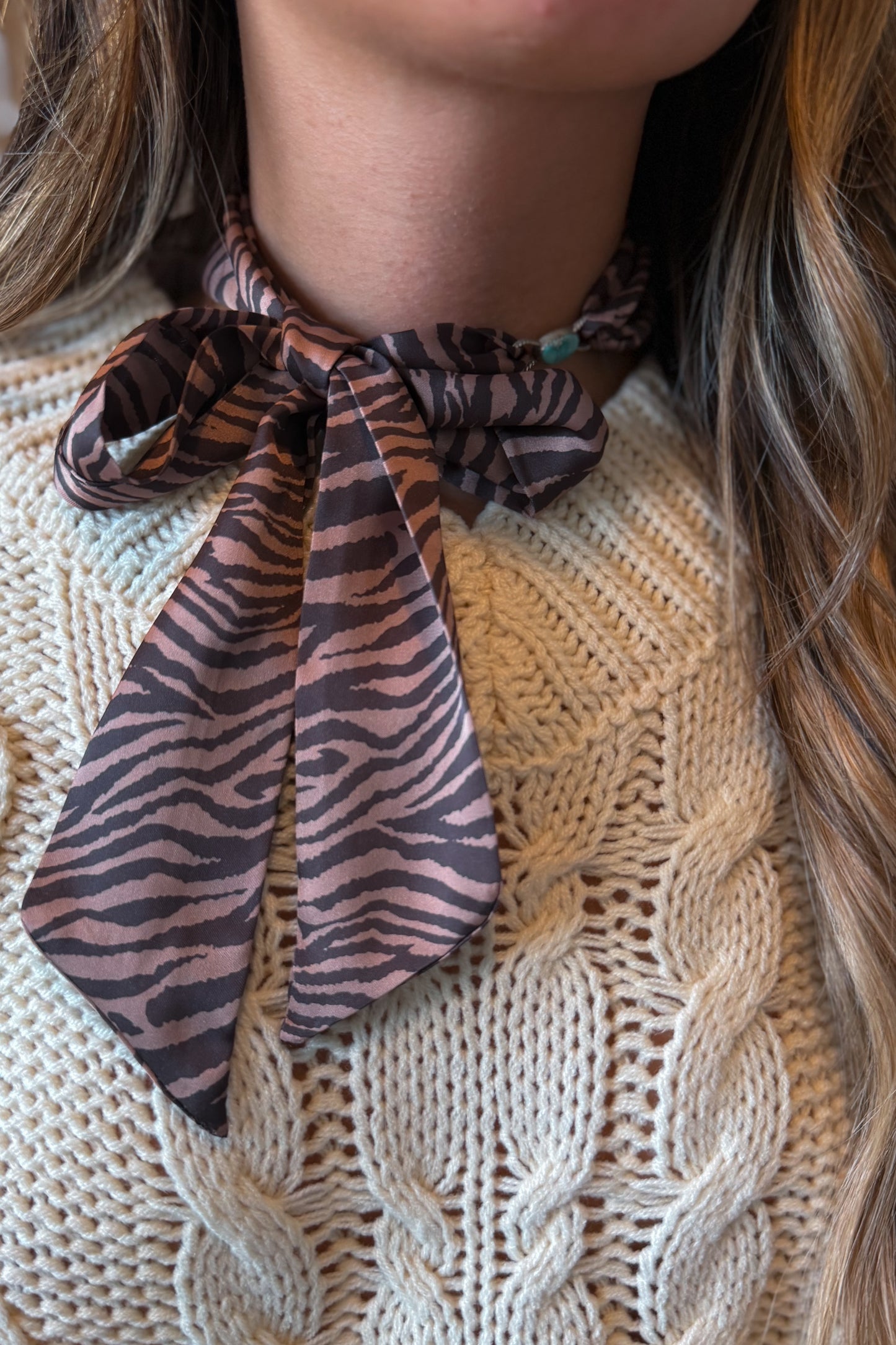 A close-up of a person wearing a brown and black zebra-patterned silk scarf tied around their neck, paired with a cream-colored cable-knit sweater.