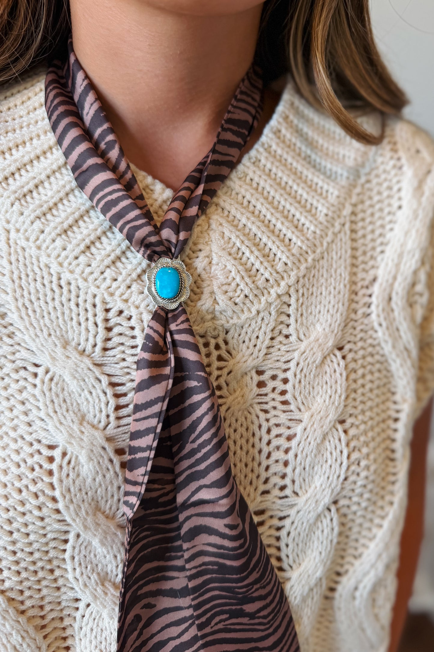 A close-up of a person wearing a cream cable-knit sweater vest over a white top, styled with a brown and black zebra-print silk necktie featuring a turquoise stone brooch.