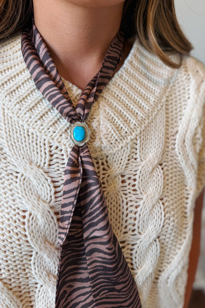 A close-up of a person wearing a cream cable-knit sweater vest over a white top, styled with a brown and black zebra-print silk necktie featuring a turquoise stone brooch.