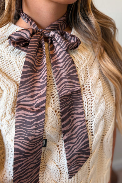 A close-up of a model wearing a brown and black zebra-print neck scarf tied in a bow over a cream cable-knit sweater.
