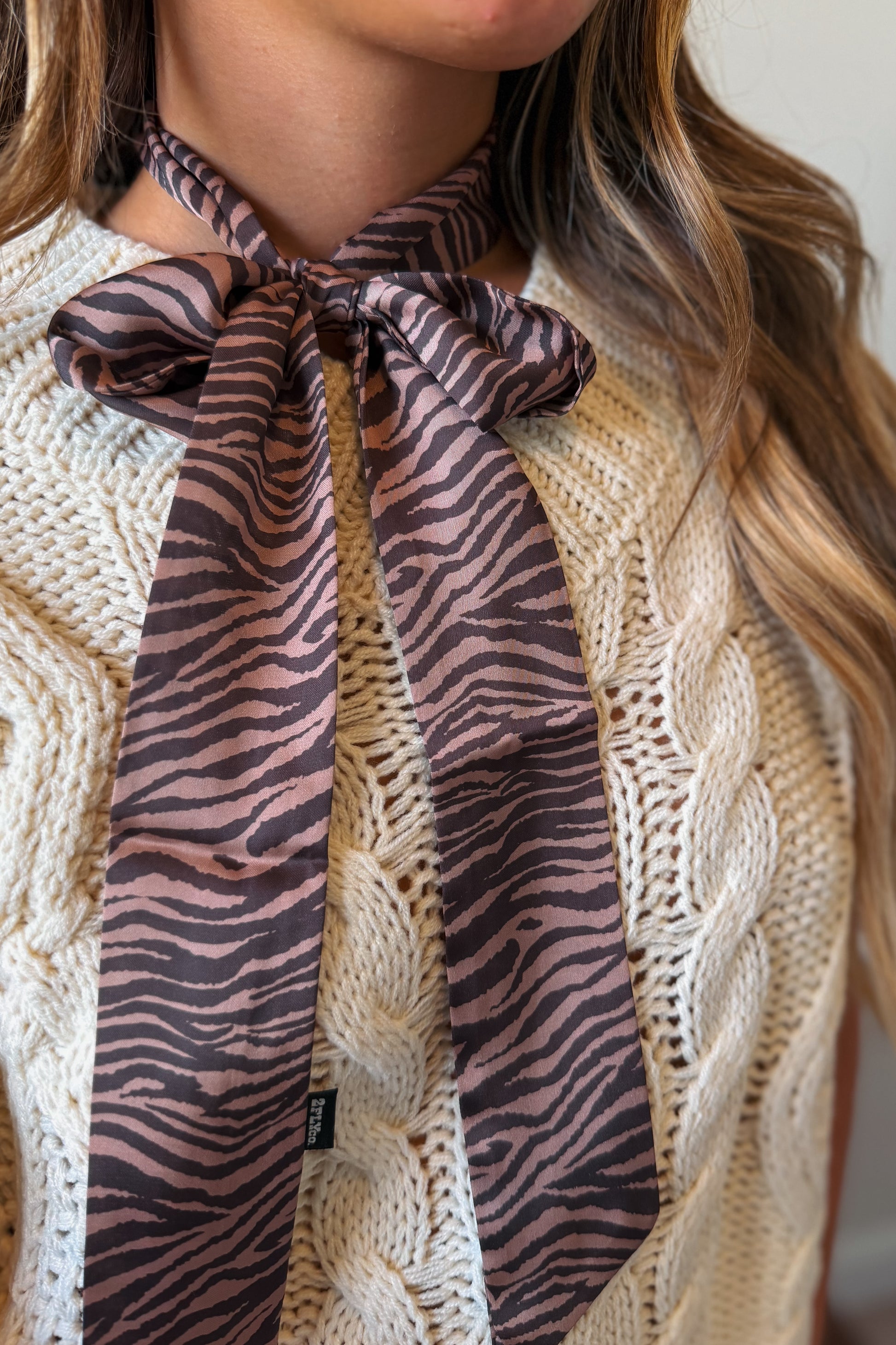 A close-up of a woman wearing a cream cable-knit sweater and a brown and black zebra-print neck scarf tied in a bow at the collar.