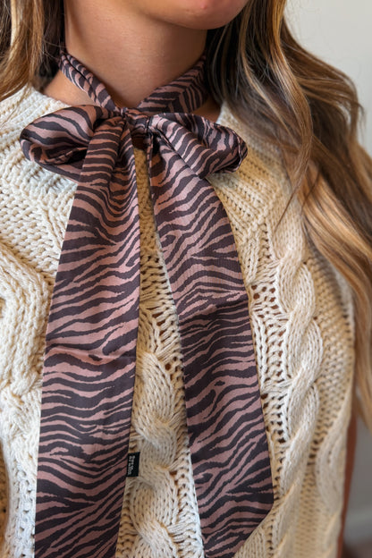 A close-up of a woman wearing a cream cable-knit sweater and a brown and black zebra-print neck scarf tied in a bow at the collar.